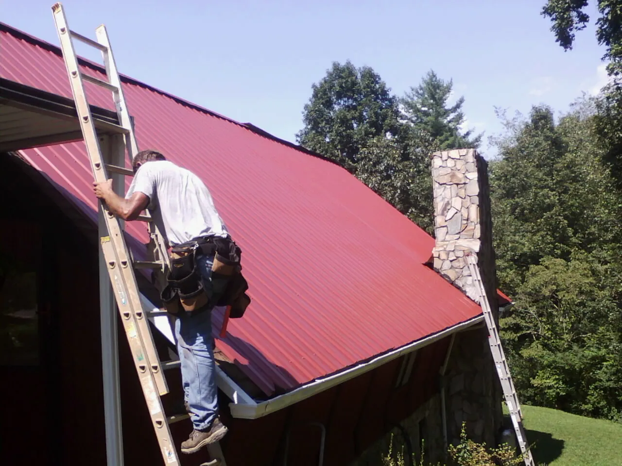 Roofer on a ladder beside a red metal roof and stone chimney — Soffit & Fascia Repair by Roofing Brothers in Sugar Grove